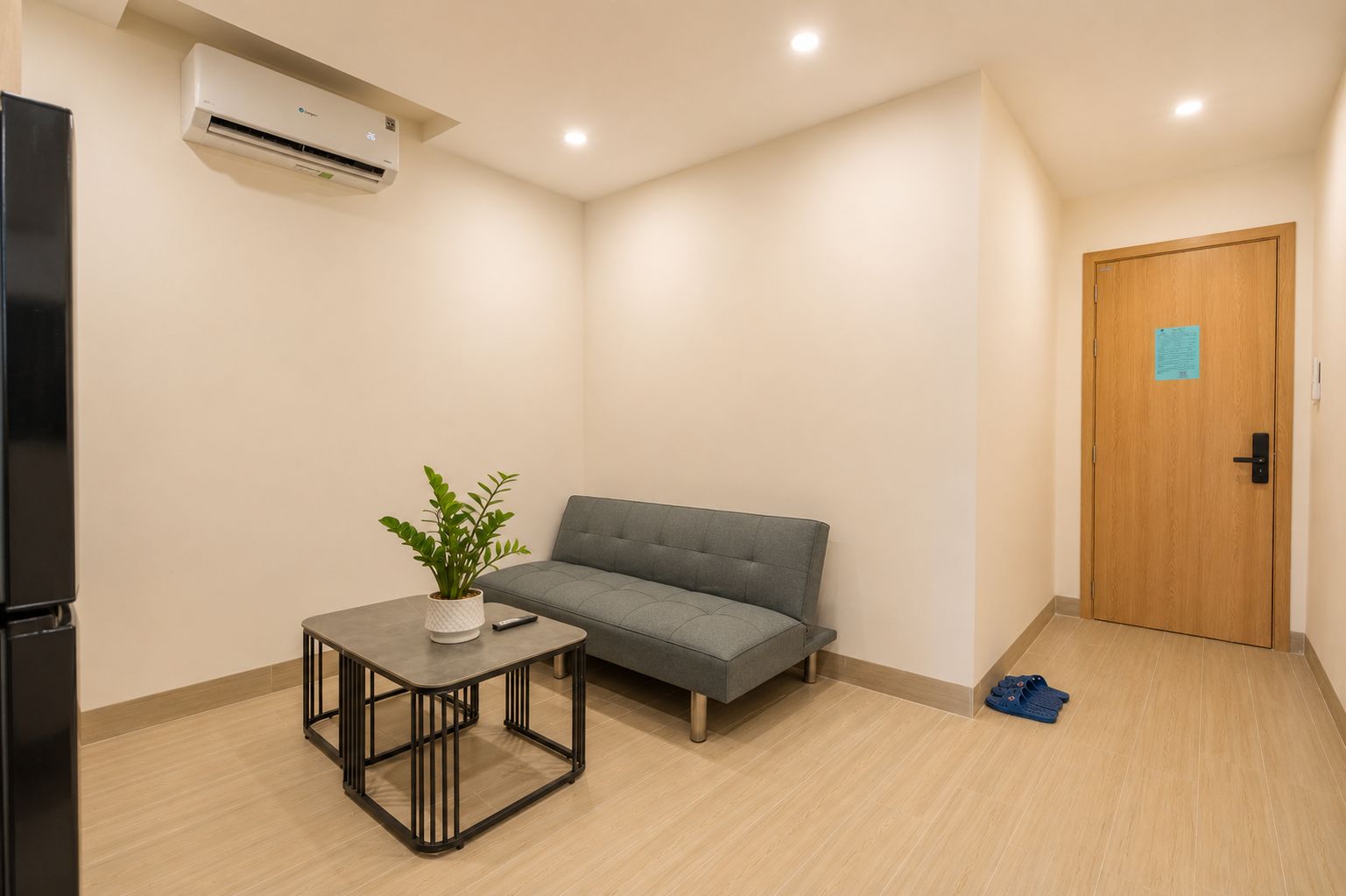 A room with a grey sofa, a small table with a plant, a black refrigerator, and a wooden door with a blue sign.