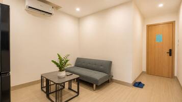A room with a grey sofa, a small table with a plant, a black refrigerator, and a wooden door with a blue sign.
