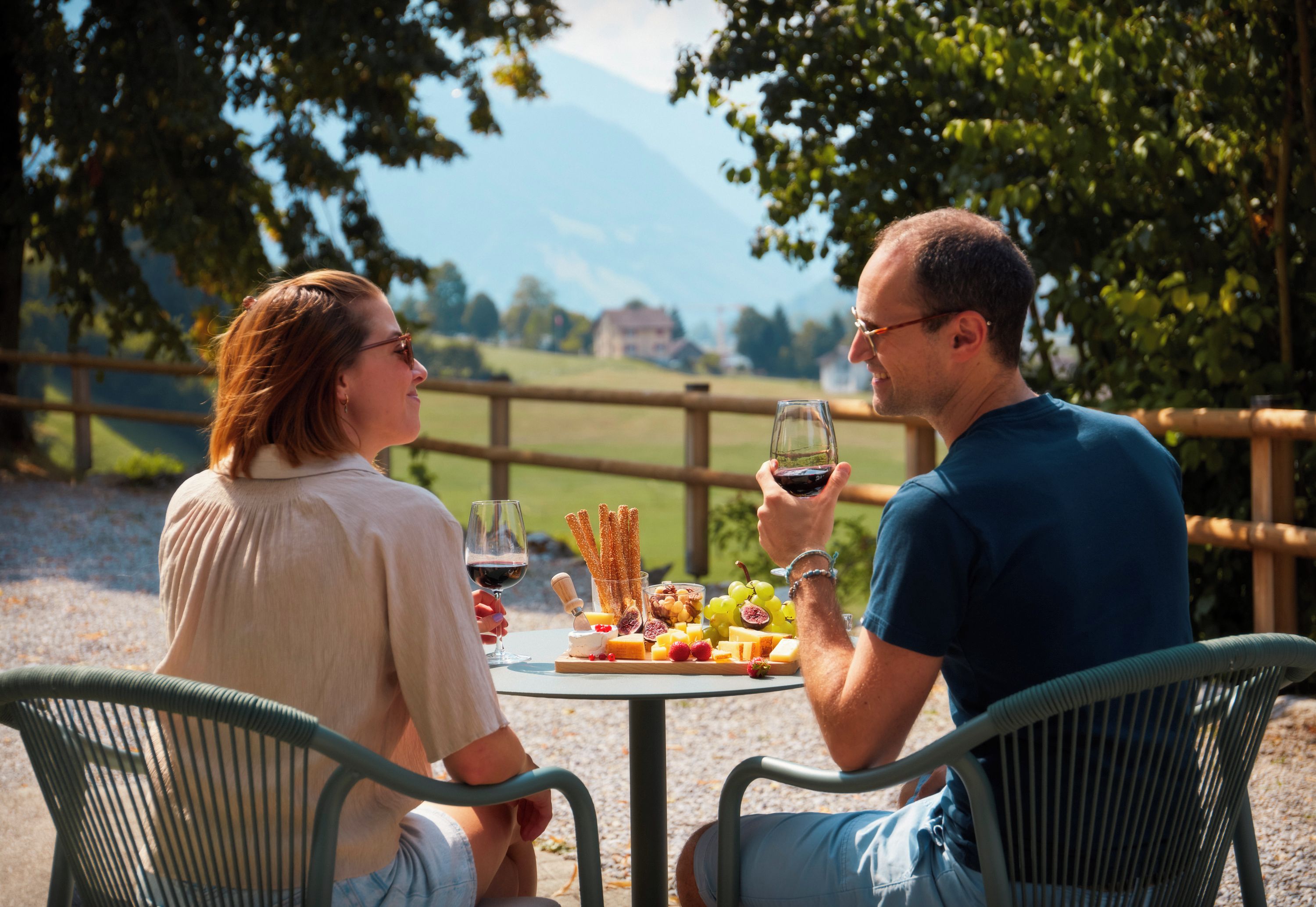 Zwei Personen sitzen an einem Tisch im Freien mit verschiedenen Speisen und Getränken, umgeben von Grünpflanzen und einem Holzzaun.