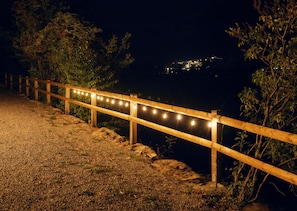A wooden pathway with string lights, leading to a body of water with a distant view of a town.