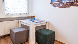 Une petite pièce avec une table blanche, deux poufs, une fenêtre avec des stores et un tableau accroché au mur.