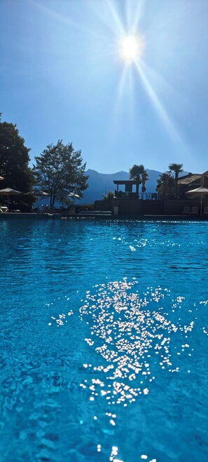 A clear blue pool with sunlight reflecting on the water, surrounded by lounge chairs and umbrellas, with mountains in the background. - Idyllic Bungalow with Lake View & Pool in Tignale (Aer)