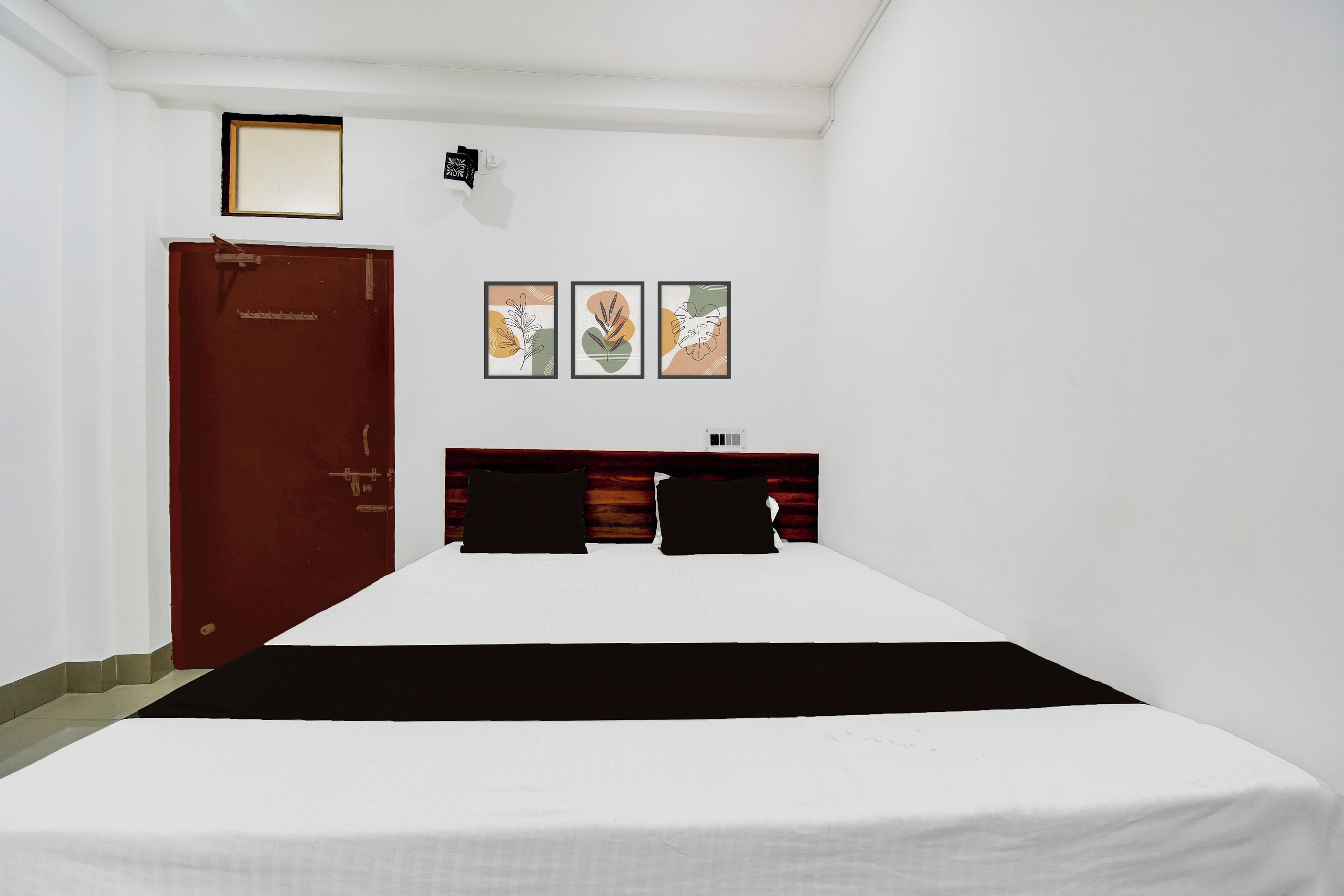 Une chambre d’hôtel avec un lit simple, une tête de lit en bois, une porte et trois œuvres d’art encadrées accrochées au mur.