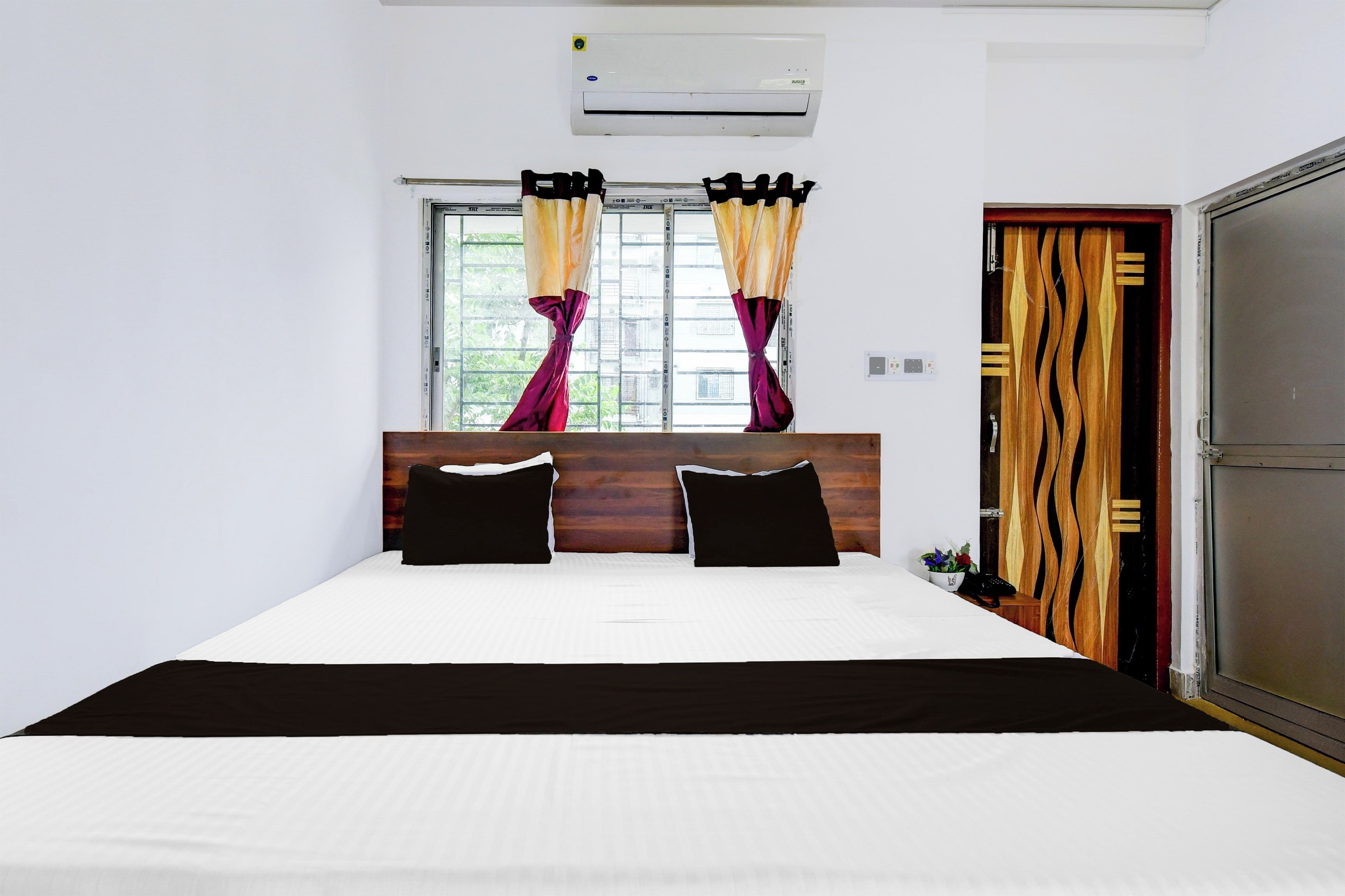 A hotel room with a large bed, a wooden headboard, an air conditioning unit, a window with curtains, and a wardrobe.