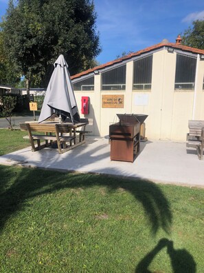 Un espacio al aire libre con mesa de picnic y parrilla, un contenedor de basura y un letrero que indica zona de barbacoa.
