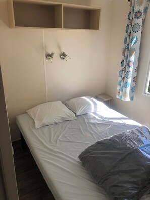 A single bed with white bedding, a small bedside table, and a wall-mounted shelf above the bed. - Mobile Home for 2–6 People (Saint-Georges-de-Didonne)