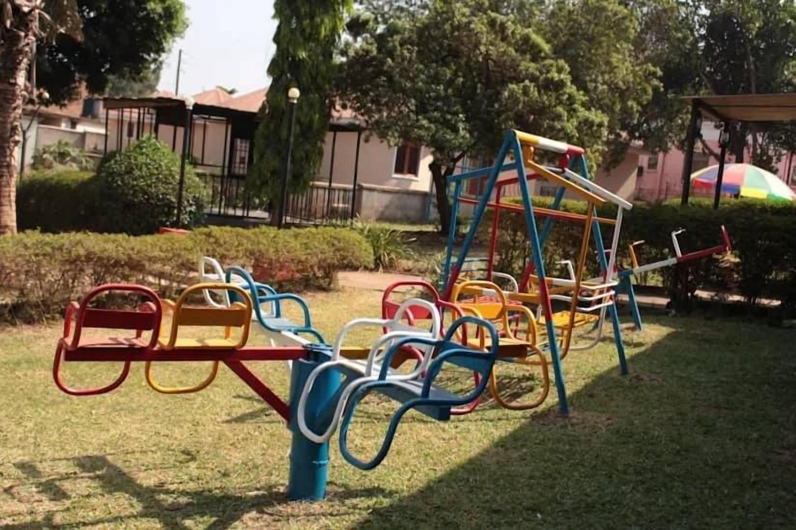 Parquinho com diversos equipamentos de exercício, incluindo balanços e um escorrega, em área gramada com árvores e edifícios ao fundo.