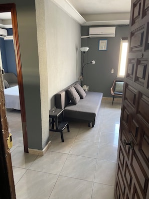 A room with a sofa, a small table, a floor lamp, and a chair. There is a door with a geometric pattern and a window with blinds. - Los Boliches one-bedroom apartment with a lift, Beach 130m, Train st. dist.0.8m (Fuengirola)