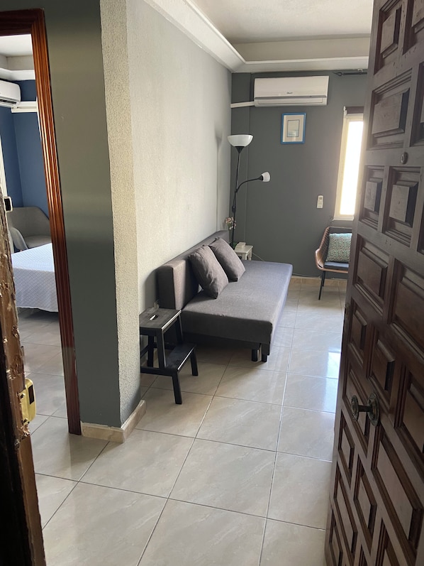 Une pièce comprenant un canapé, une petite table, un lampadaire et une chaise. On y trouve une porte ornée d’un motif géométrique et une fenêtre avec des stores.
