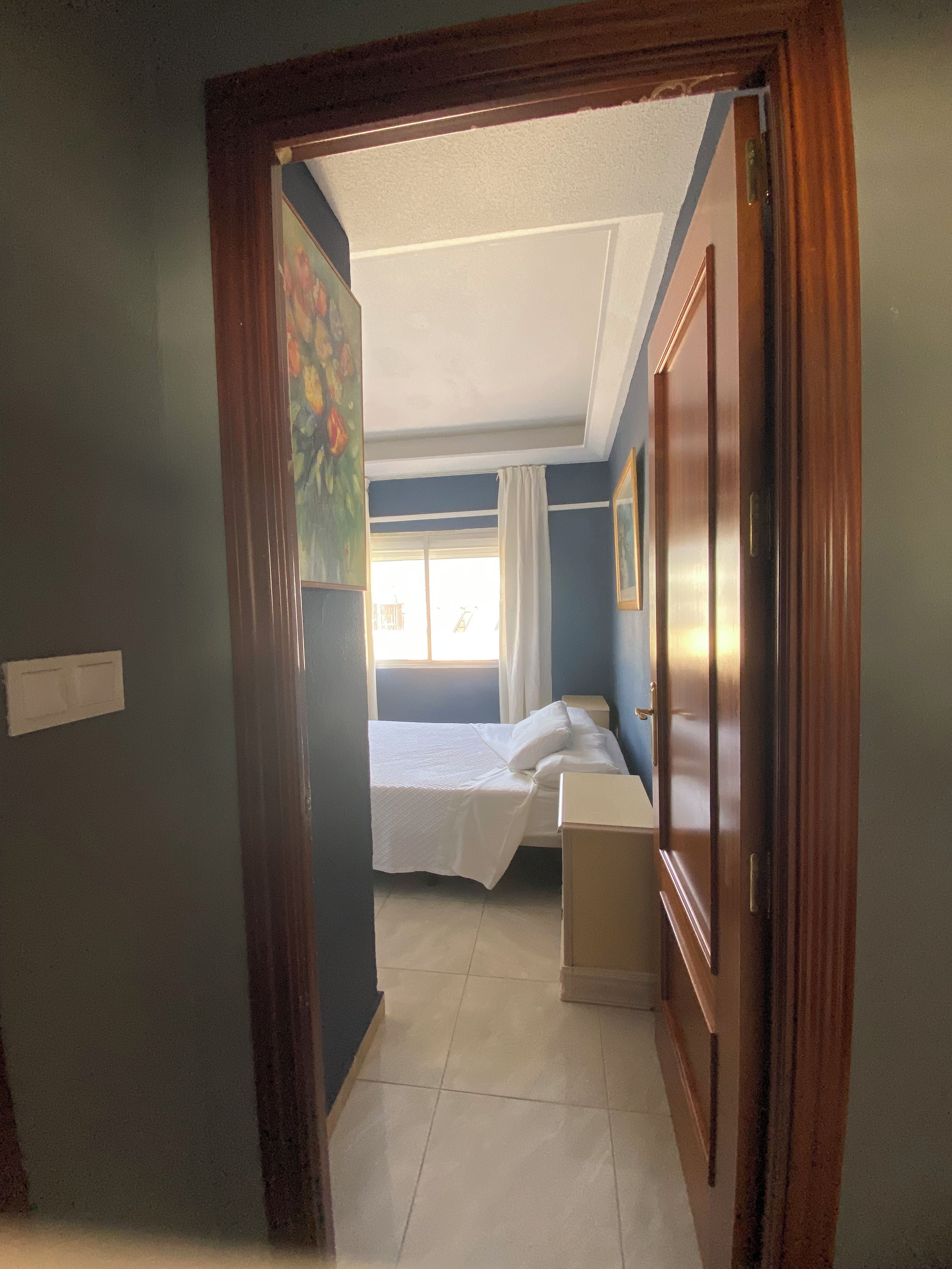 A room with a bed, a bedside table, a window with curtains, and a door.