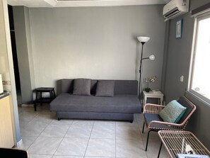 Un salon moderne avec un canapé gris, une petite table d’appoint, un lampadaire et un fauteuil en osier.