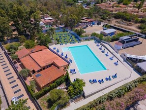 Vista aérea de un hotel con una gran piscina rodeada de sillas de descanso y sombrillas, junto a un edificio con techo de tejas rojas.