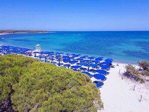 Una playa con numerosas sombrillas azules y sillas reclinables blancas, una torre de salvavidas y una vista despejada del mar.