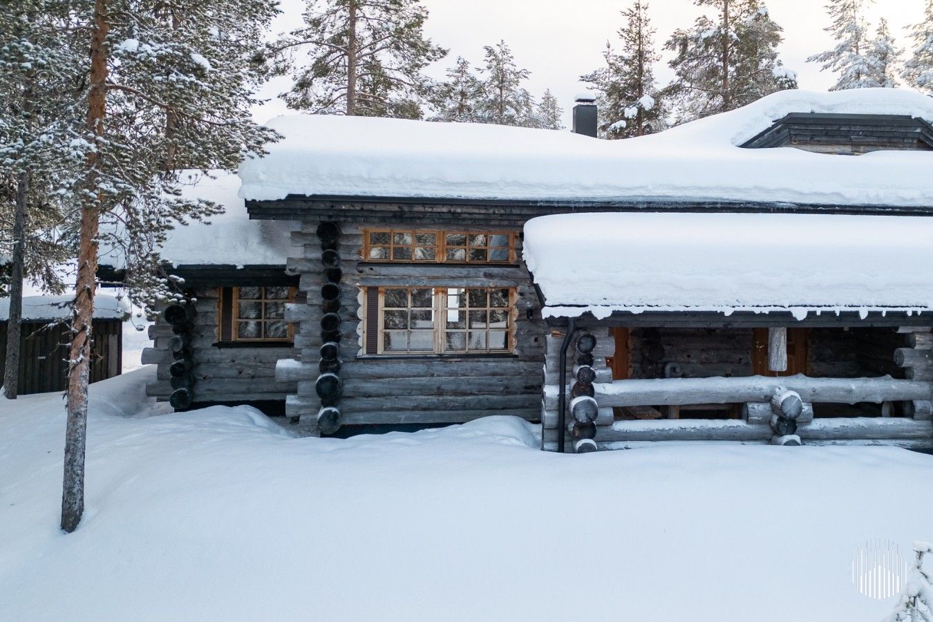 Une cabane en rondins dont le toit et les fenêtres sont recouverts de neige, entourée d’arbres.