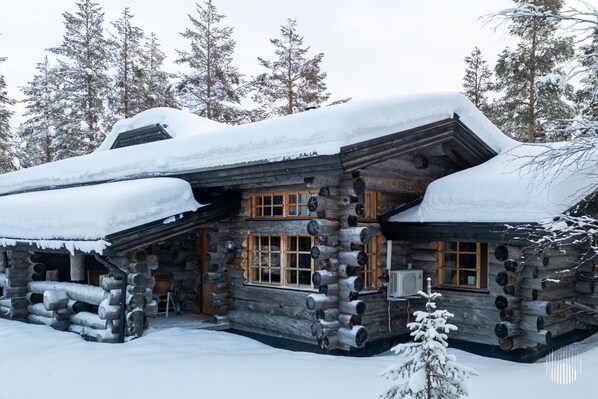 A log cabin with a snow-covered roof, wooden windows, and a log exterior.