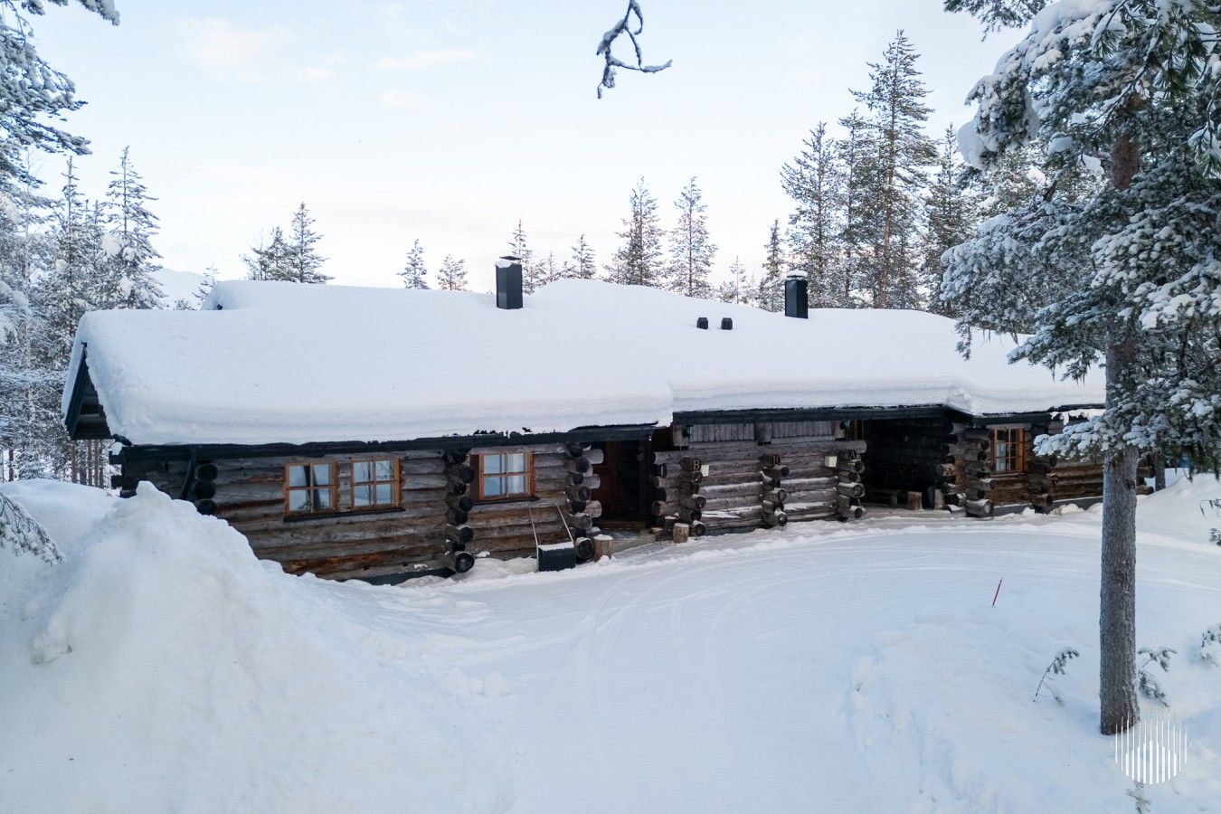 Une cabane en rondins dont le toit est recouvert de neige, entourée d’arbres également couverts de neige.