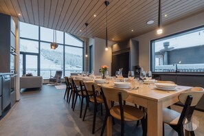 A modern dining area with a long table set for six, large windows offering a view of a snowy landscape, and a kitchen area with modern appliances. - Levi West Peak View by Holiday in Lapland (Levi)
