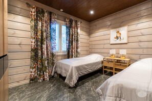 A room with two beds, wooden walls, a window with curtains, a desk with lamps, and a framed picture on the wall.