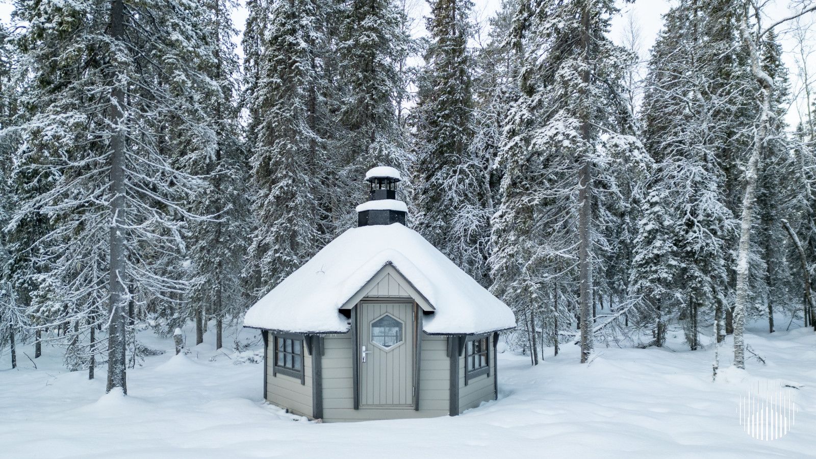 En liten, snötäckt stuga med ett brant tak, omgiven av höga träd som är täckta av snö.