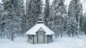 A small, snow-covered cabin with a steep roof, surrounded by tall, snow-laden trees.