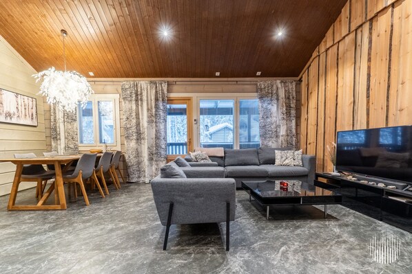 A modern living room with a sofa, coffee table, and TV. There is a dining area with chairs and a table. The room has wooden walls and a ceiling with recessed lighting.