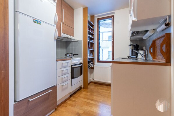 A compact kitchen with a refrigerator, oven, and stove, wooden cabinets, and a window with blinds.