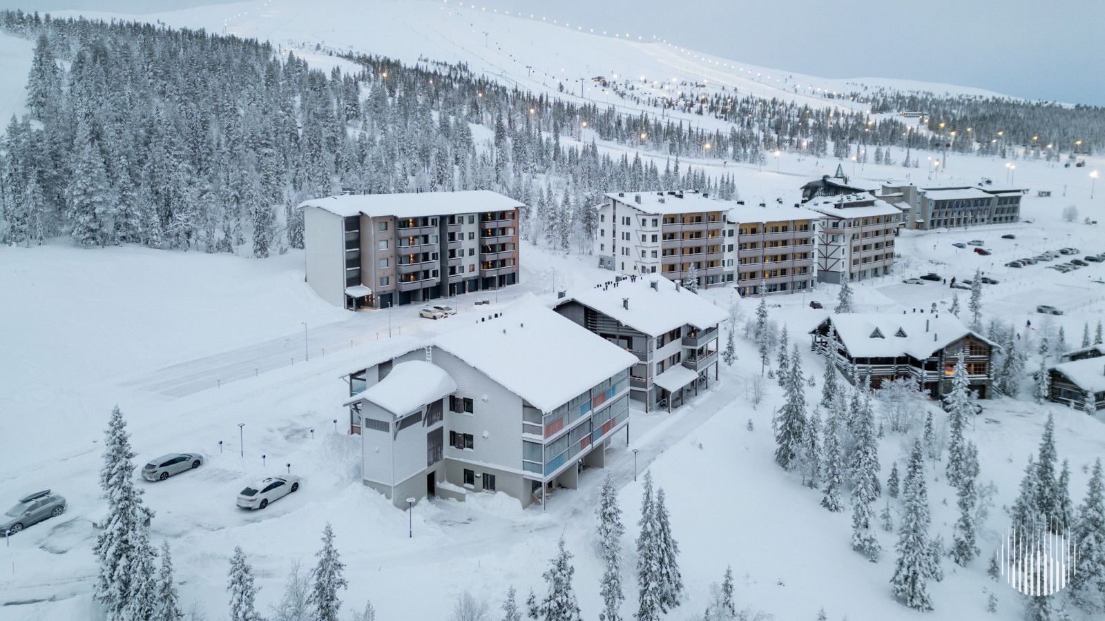 Una località sciistica innevata con diversi edifici, tra cui un grande albergo con numerosi piani e balconi, circondata da alberi e una pista da sci sullo sfondo.