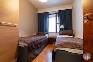 Una habitación pequeña y funcional con dos camas, una ventana con cortinas, una mesita de noche con lámpara y una puerta de madera.