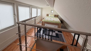 A bedroom with a bed, a window with blinds, a wooden floor, and a staircase with a metal railing. - Feldblick Rastede Vacation Rental – Covered Patio, Air Conditioning, Dogs Welcome (Rastede)