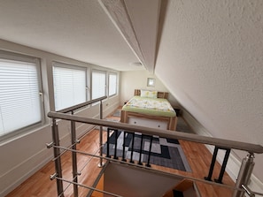 Une chambre à coucher avec un lit, une fenêtre avec des stores, un plancher en bois et un escalier doté d’une rampe en métal.