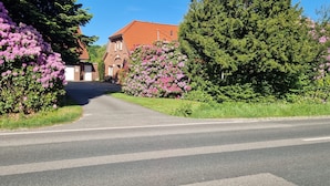 A road leading to a building with a red roof, surrounded by lush greenery and flowering bushes. - Feldblick Rastede Vacation Rental – Covered Patio, Air Conditioning, Dogs Welcome (Rastede)