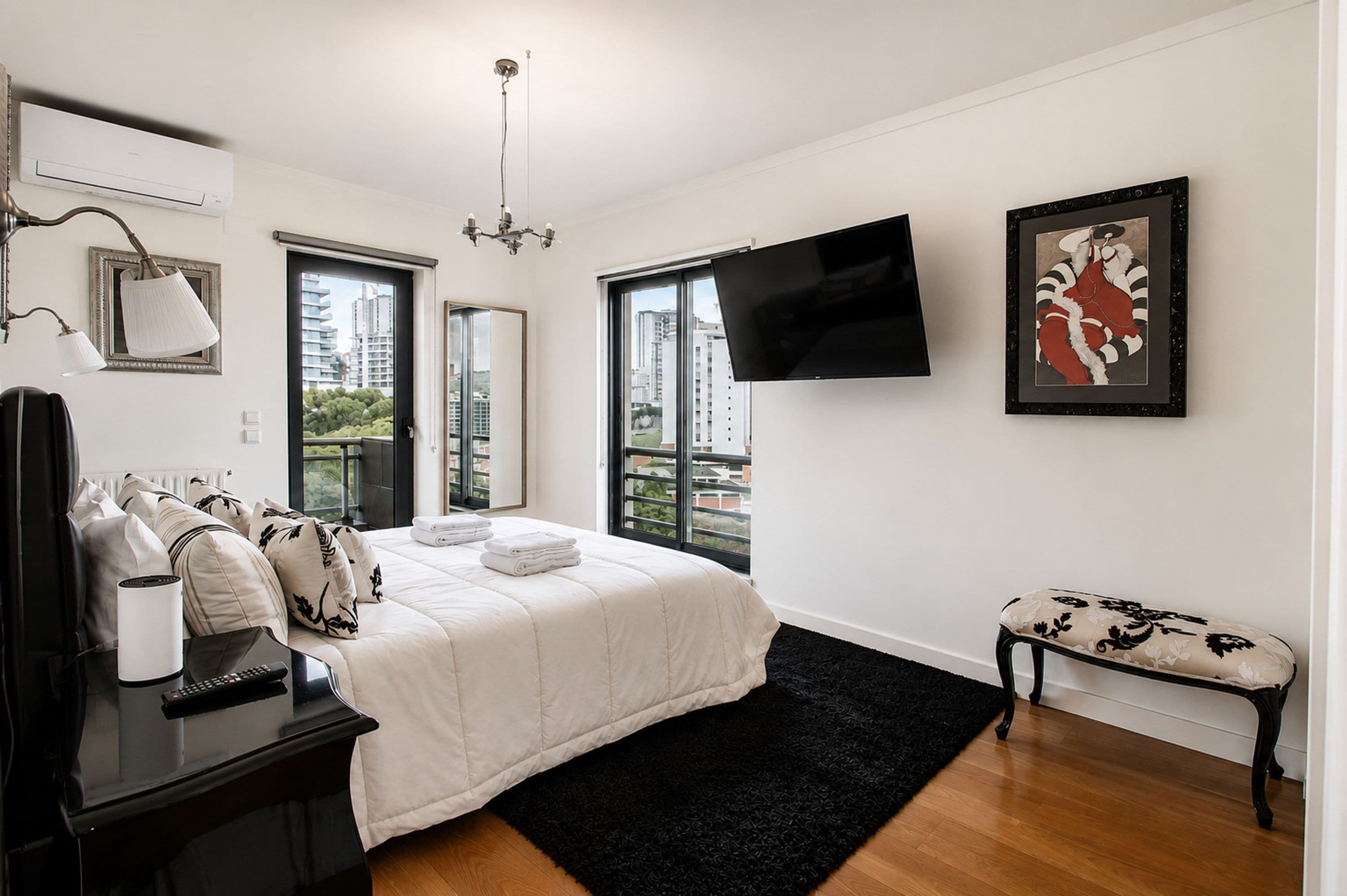 A modern bedroom with a bed, a desk, a chair, a TV, and a window with a view of buildings.
