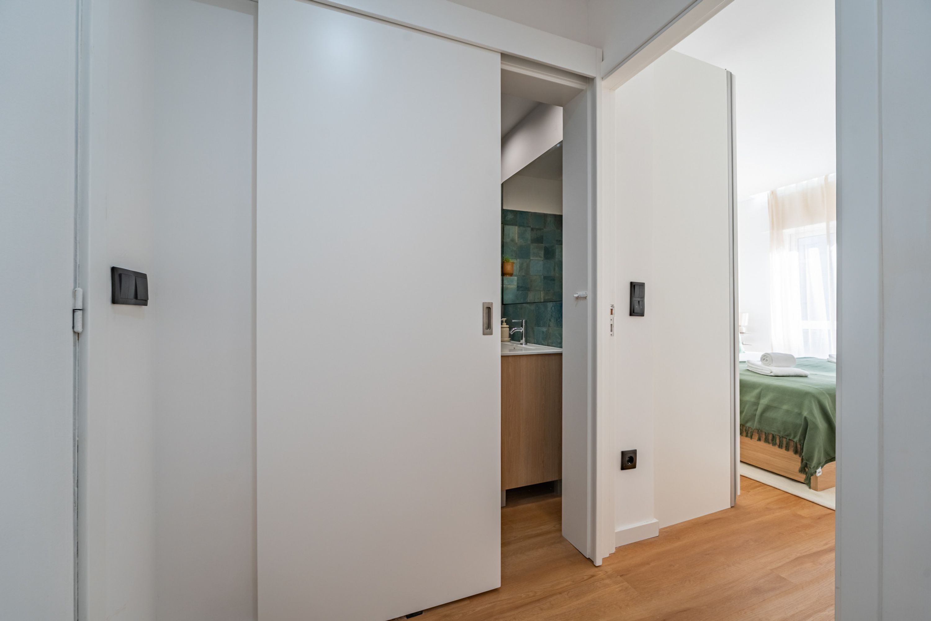 Habitación de hotel con piso de madera, cubrecama verde y baño con pared de azulejos y lavamanos.