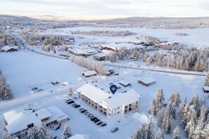 Ett snötäckt landskap med ett stort byggnadskomplex, flera mindre byggnader och en parkeringsplats.
