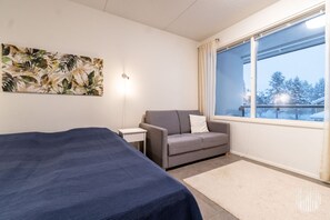Ein Zimmer mit einem Bett, einer Couch, einem kleinen Tisch, einer Wandleuchte und einem Fenster mit Blick auf Bäume.
