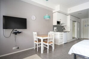 Una habitación moderna con televisión de pantalla plana en la pared, una mesa de comedor con sillas, una zona de cocina con microondas y una cama con cubierta blanca.
