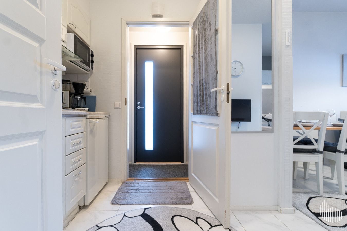 A narrow hallway with a modern kitchen on one side, a dining area with a table and chairs on the other, and a door with a vertical light strip.