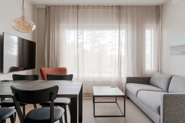 Ein modernes Wohnzimmer mit Essbereich, einer Couch, einem Fernseher und einem großen Fenster mit transparenten Vorhängen.