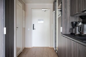 Un couloir mène à une porte blanche dotée d’une petite fenêtre, le sol est en bois et on y trouve un coin cuisine avec une cuisinière et une machine à café.