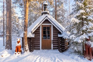 Une cabane en rondins avec une porte en bois et une cheminée, entourée d’arbres couverts de neige.