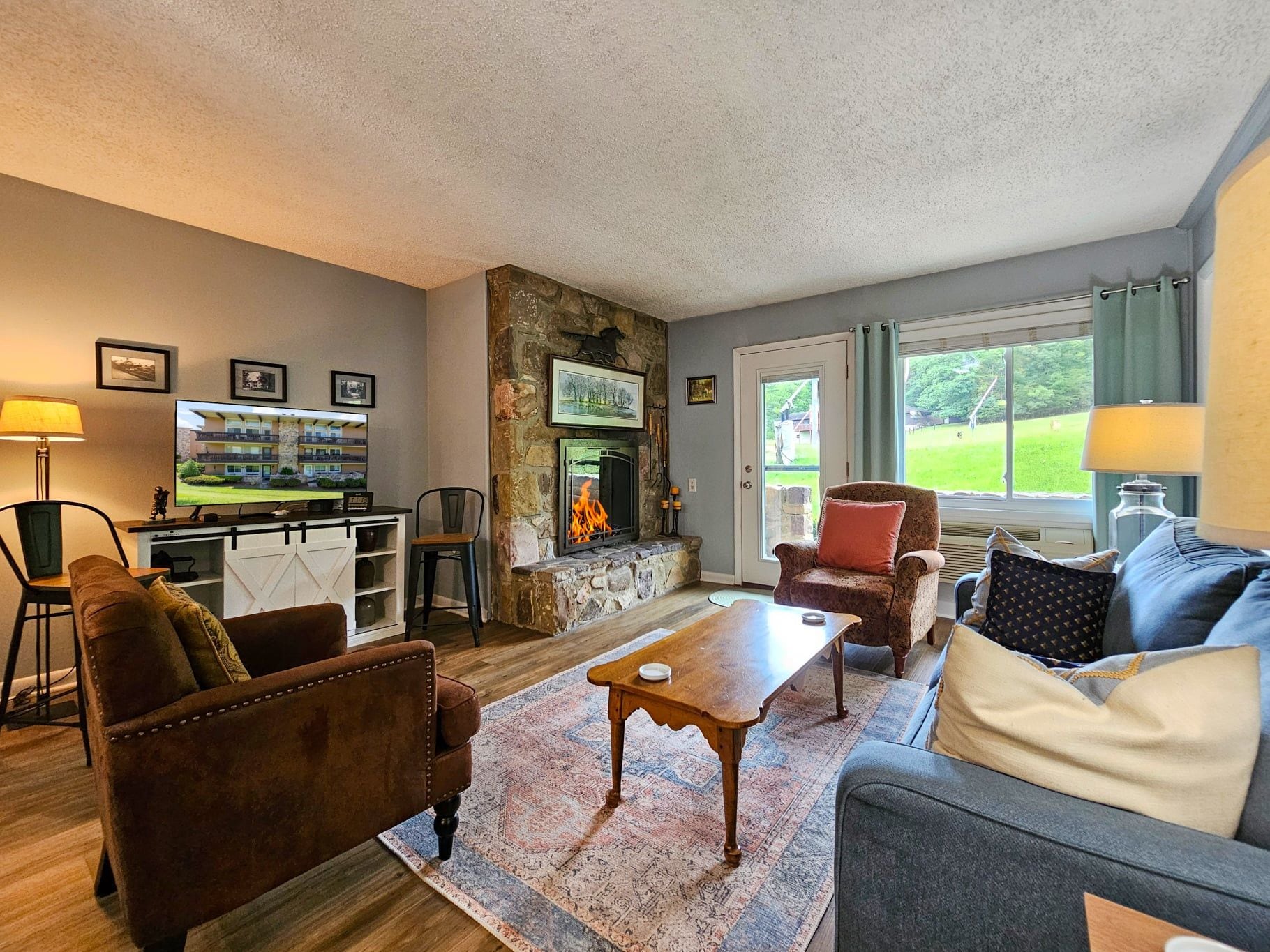 A cozy living room with a stone fireplace, comfortable seating, a television, and a view of the outdoors.
