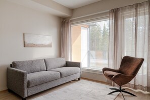 Ein Wohnzimmer mit einem grauen Sofa, einem braunen Sessel, einem Bild an der Wand und einem Fenster mit Vorhängen.