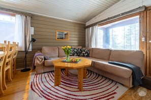 A cozy living room with a sofa, a wooden coffee table with fruits and flowers, a red and white striped rug, and a large window with curtains. - Kelokaltiokylä 29 by Holiday in Lapland (Ruka)