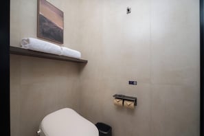 Et badeværelse med toilet, håndklædestang med håndklæde, toiletpapirholder og vægmonteret lysarmatur.