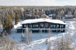 A large, two-story building with a flat roof, surrounded by snow-covered trees and a forested area. - Metsonlinna 205 by Holiday in Lapland (Levi)