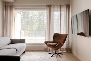 Ein Zimmer mit grauem Sofa, braunem Sessel, großem Fenster mit Jalousien und einem Wand-Fernseher.