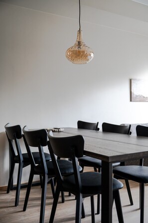 Ein Essbereich mit Holztisch und schwarzen Stühlen, einer hängenden Leuchte sowie einer schlichten Wand mit einem gerahmten Bild.