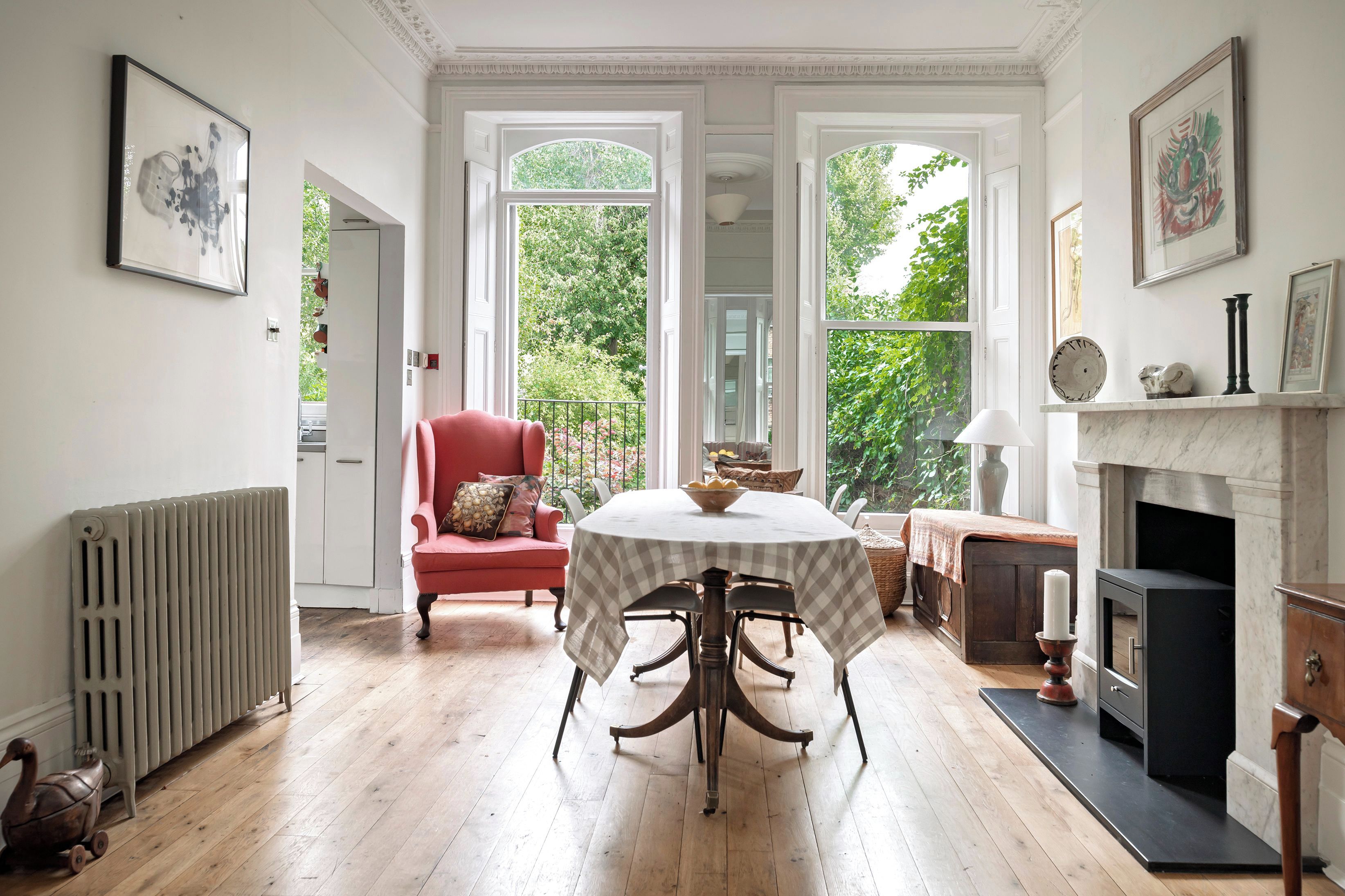 A room with a dining table, a red armchair, a fireplace, and large windows with a view of greenery.