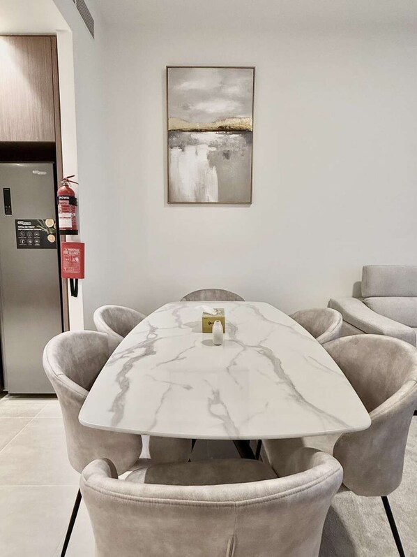 A dining area with a marble table, cushioned chairs, a fire extinguisher, a fire blanket, and a framed abstract painting on the wall.