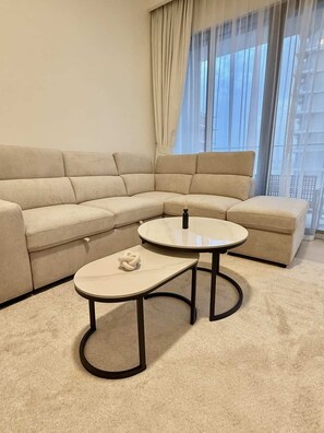 A corner living room with a beige sectional sofa, two round tables, and a view of a building through sheer curtains.